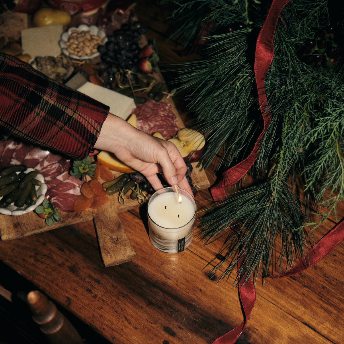 A hand wearing a red plaid sleeve lights a Ranger Station Cowboy Christmas candle on a rustic wooden table surrounded by holiday greenery, pine branches, and a charcuterie board.