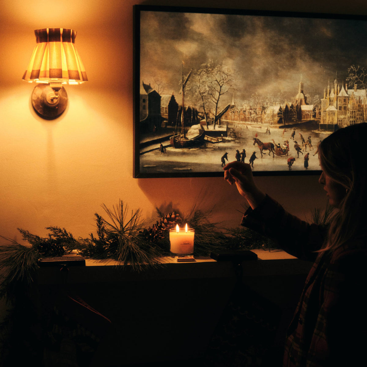 A person in silhouette stands beside a lit Ranger Station Cowboy Christmas candle and a framed winter cityscape painting on a warm wall, with a glowing sconce above.