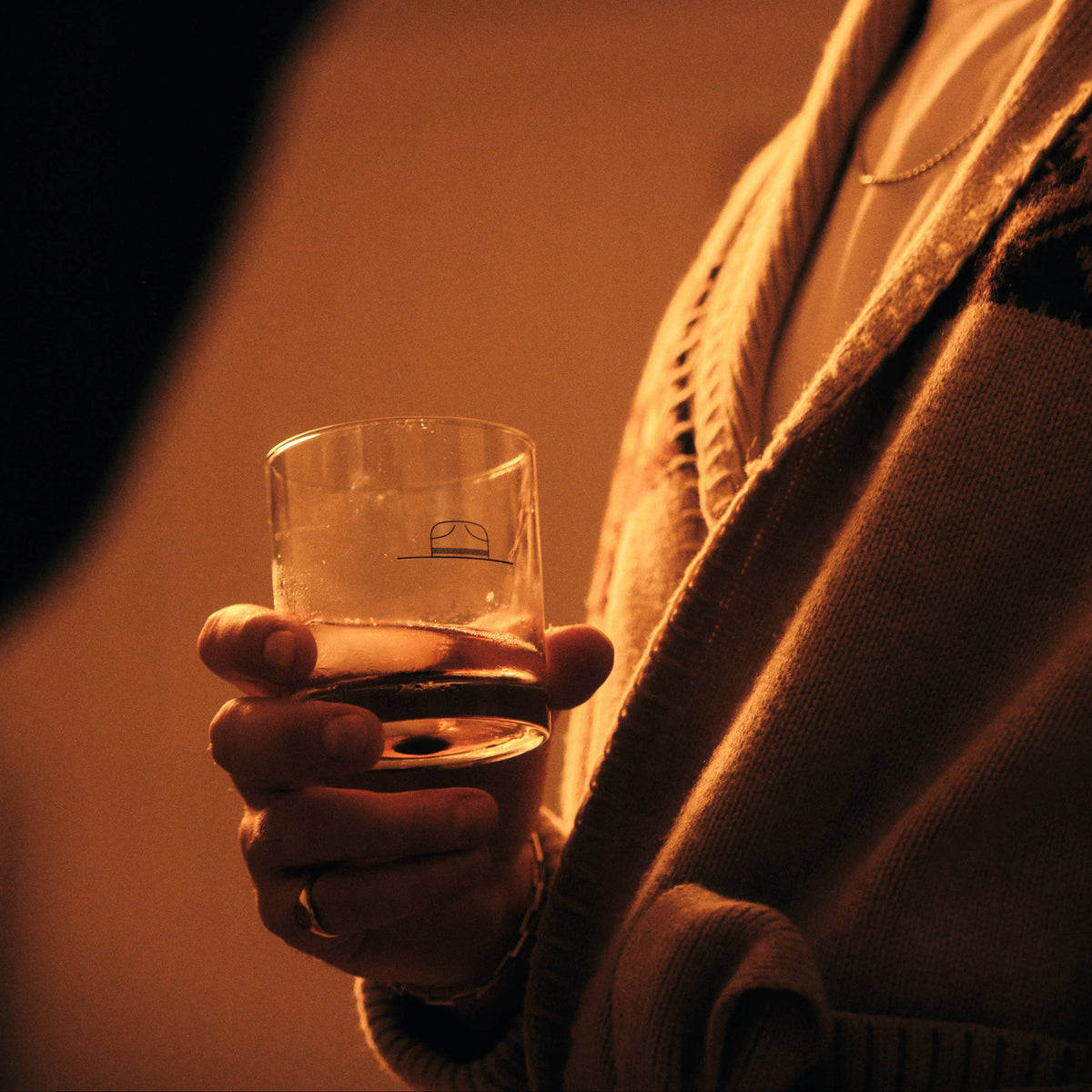 A warm, low-light lifestyle shot of a man in a beige knit cardigan and hat holding a clear glass tumbler with a small logo, set against a dark, shadowed background.