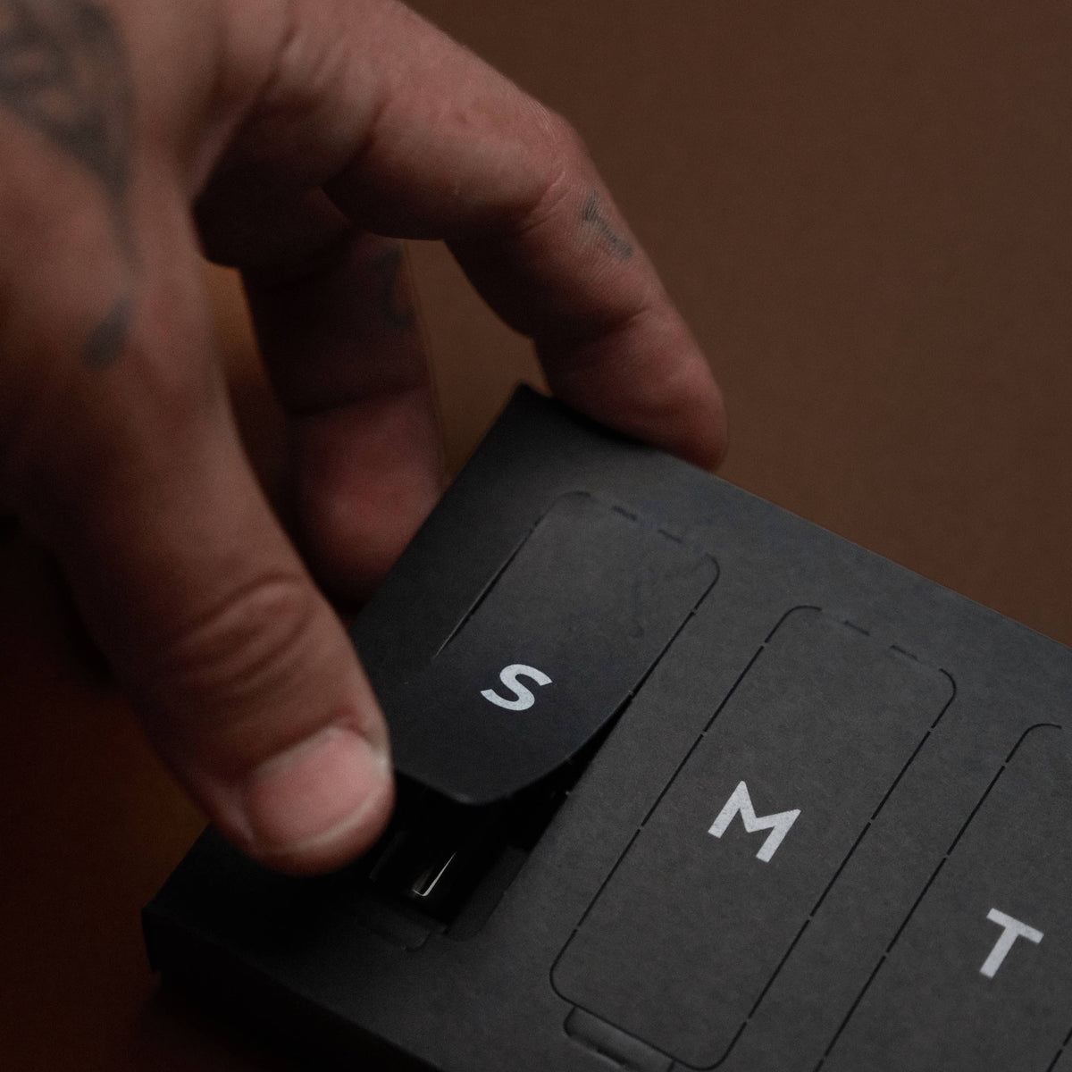 A close-up of a hand with tattoos pressing the 'S' size button on a black textured package of Ranger Station Fragrance, with 'M' and 'T' buttons visible behind it against a brown background.