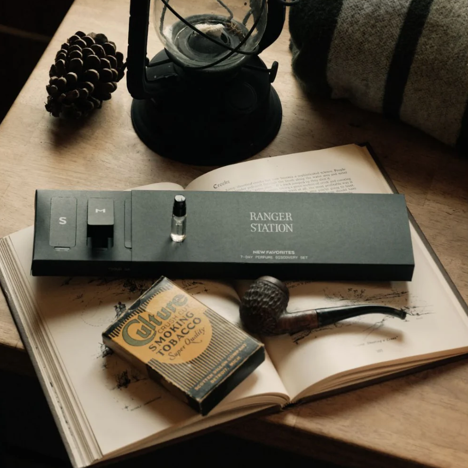A dark gray rectangular package of fragrances labeled 'RANGER STATION' rests on an open book, alongside a vintage pack of Glue Smoking Tobacco, a pipe, a pinecone, and a lantern on a wooden surface.