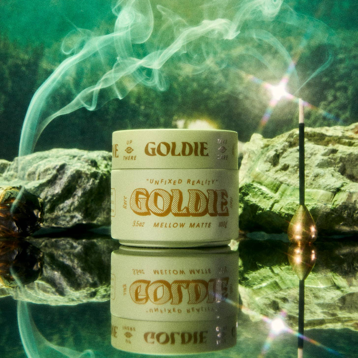 A jar of Goldie Mellow Matte Pomade sits on a reflective surface with a smoking incense stick and green mist in the background.