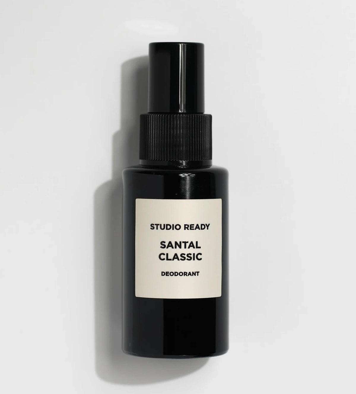 A black spray bottle of deodorant with a white label reading 'STUDIO READY SANTAL CLASSIC DEODORANT', photographed on a light gray background with a soft shadow.