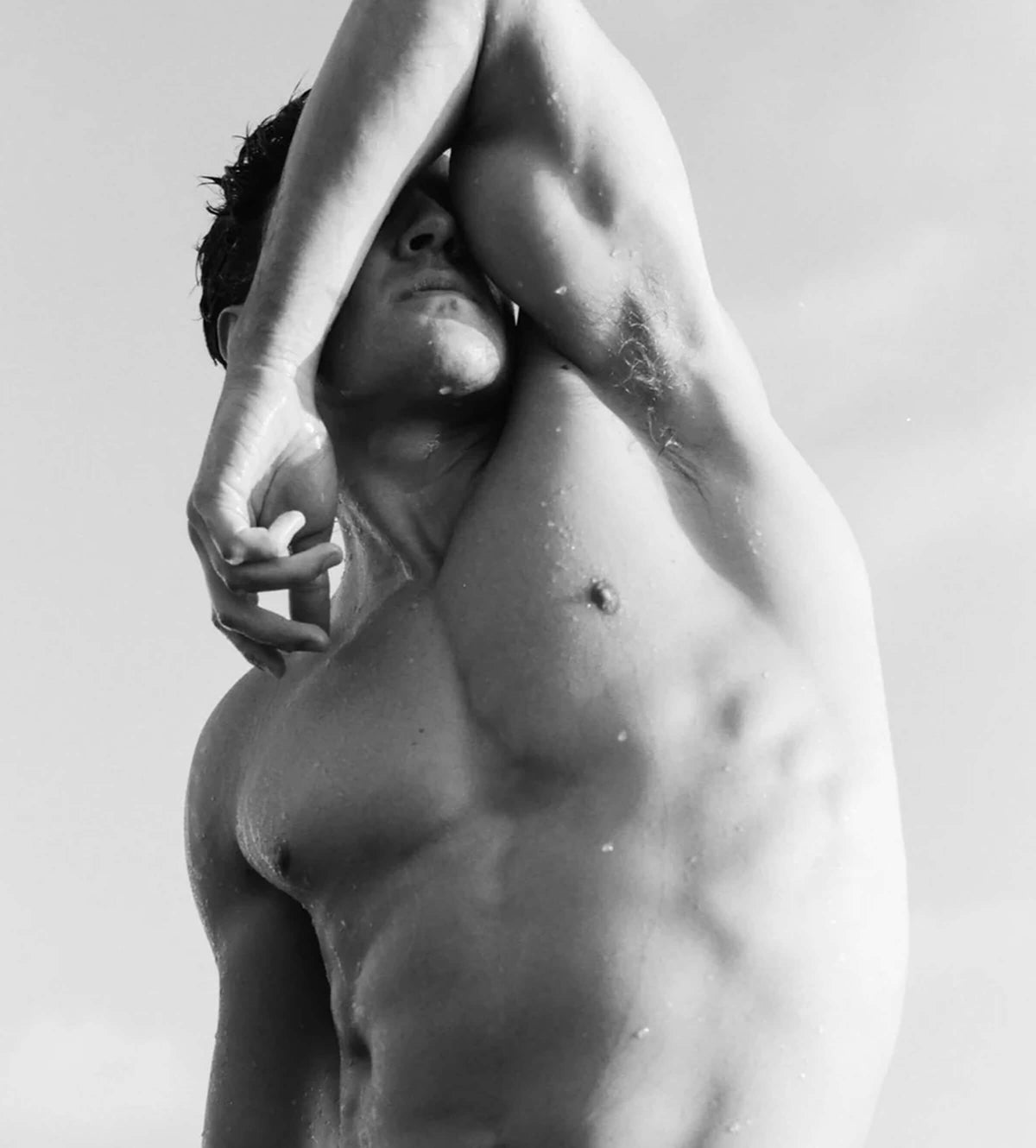 Black and white close-up photograph of a muscular man with his arms raised over his head, set against a bright sky.