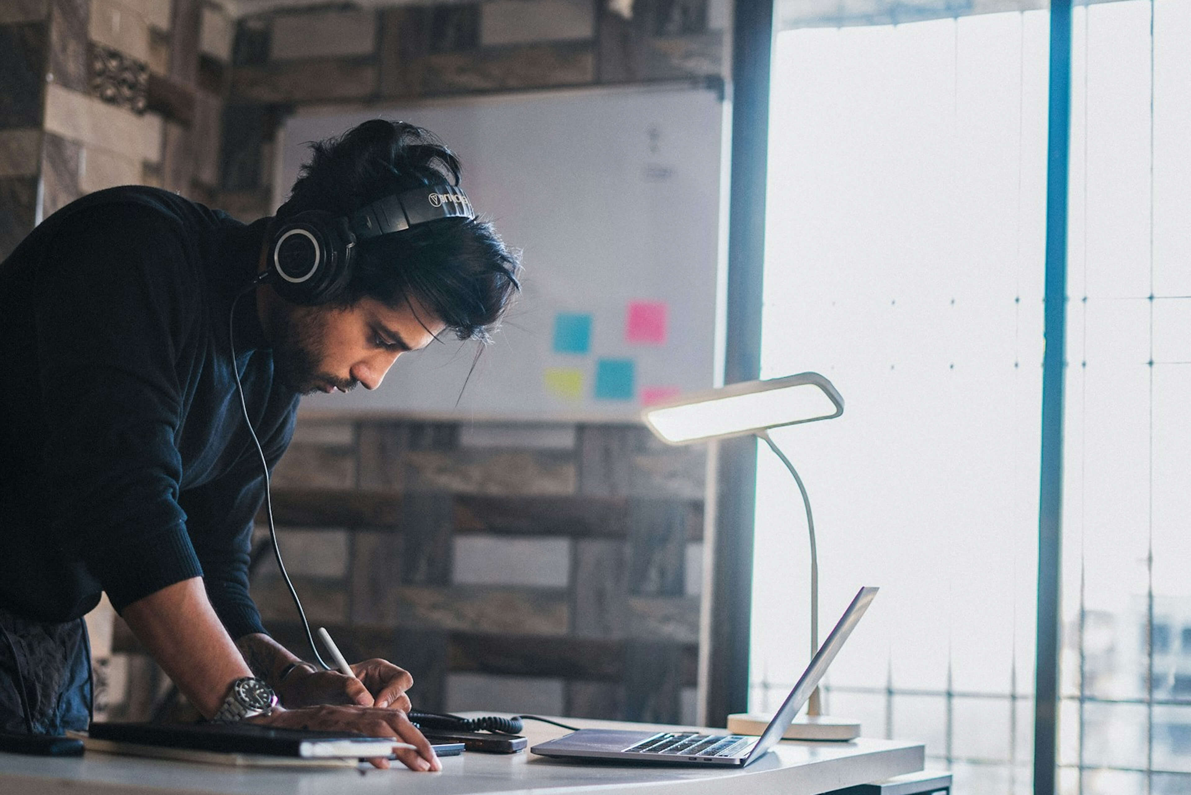 Person wearing headphones and working on a laptop with a desk lamp in a room.