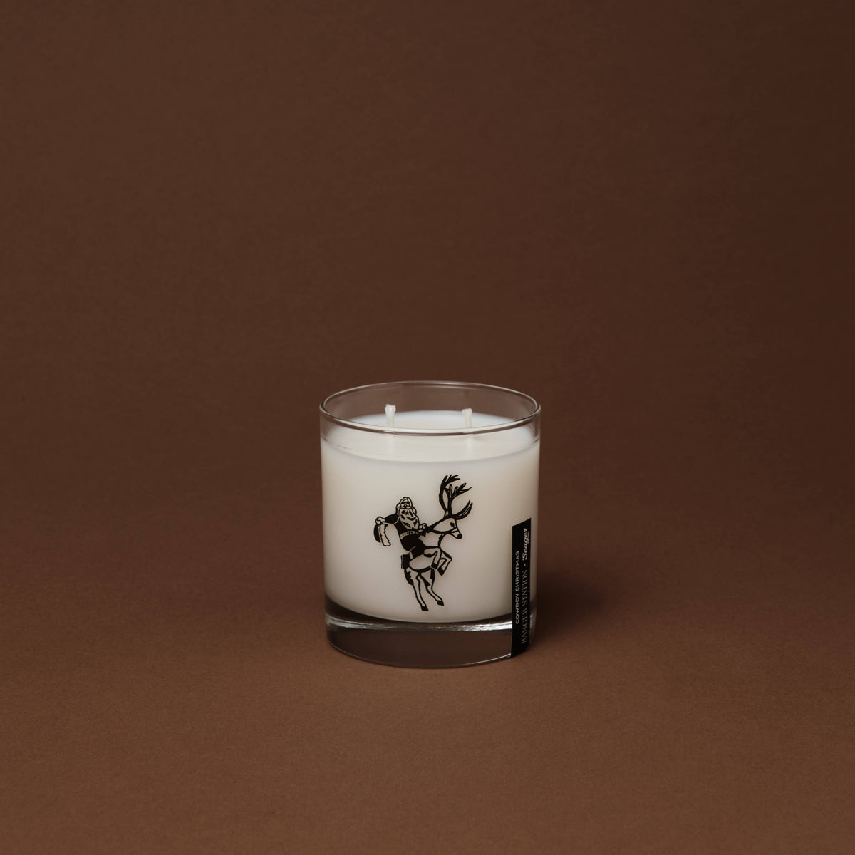A white scented Ranger Station Cowboy Christmas candle in a clear glass jar with a black line-art illustration of a deer on the front, set against a solid dark brown background.