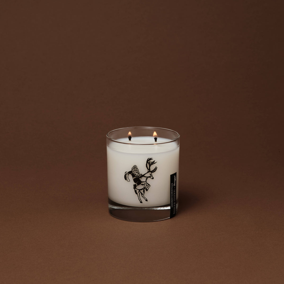 A double-wick Ranger Station Cowboy Christmas scented candle in a clear glass jar with white wax, featuring a black line-art illustration of a deer on the front, set against a solid dark brown background.