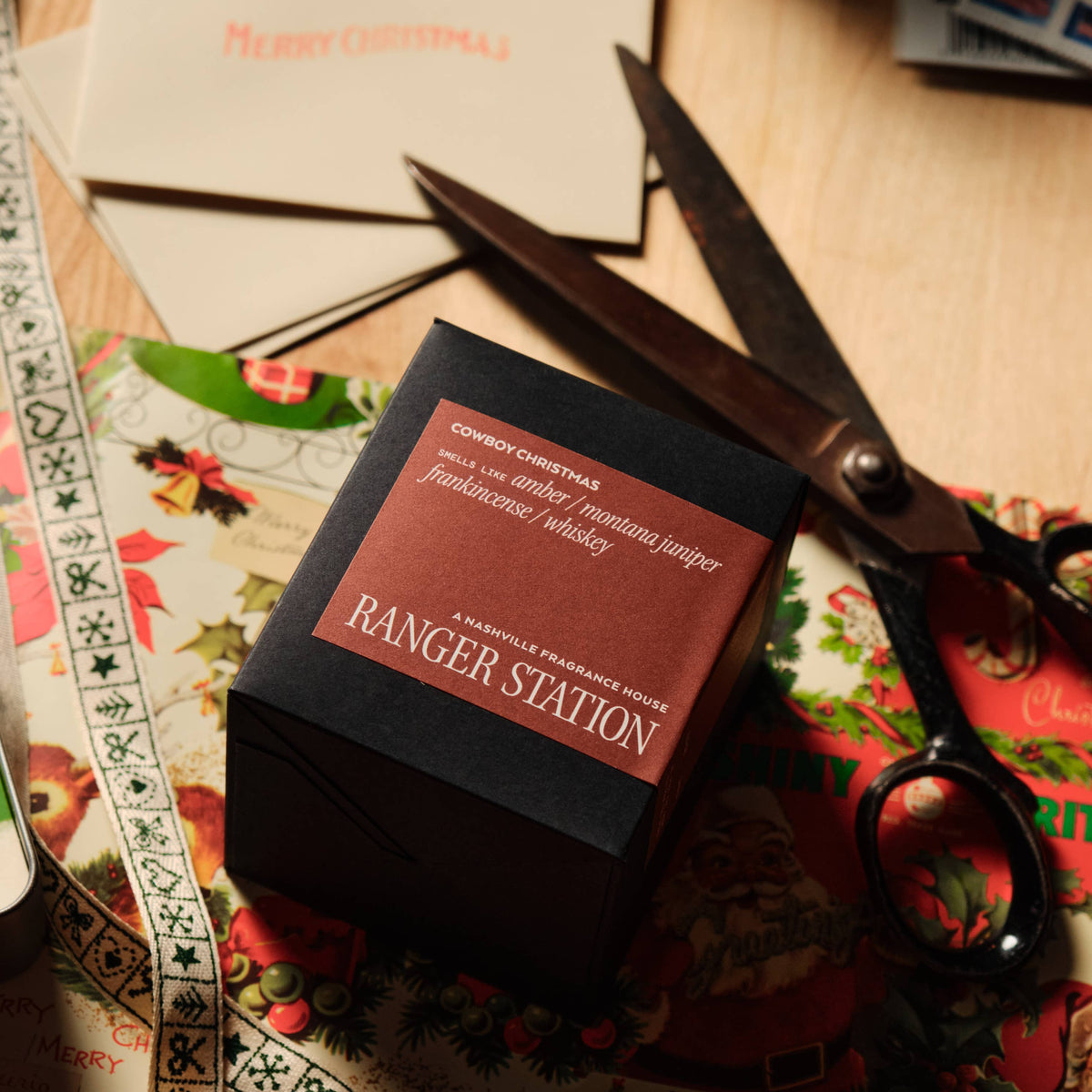 A small black gift box with a red label reading 'RANGER STATION' sits on a tabletop surrounded by Christmas cards, scissors, and patterned paper.