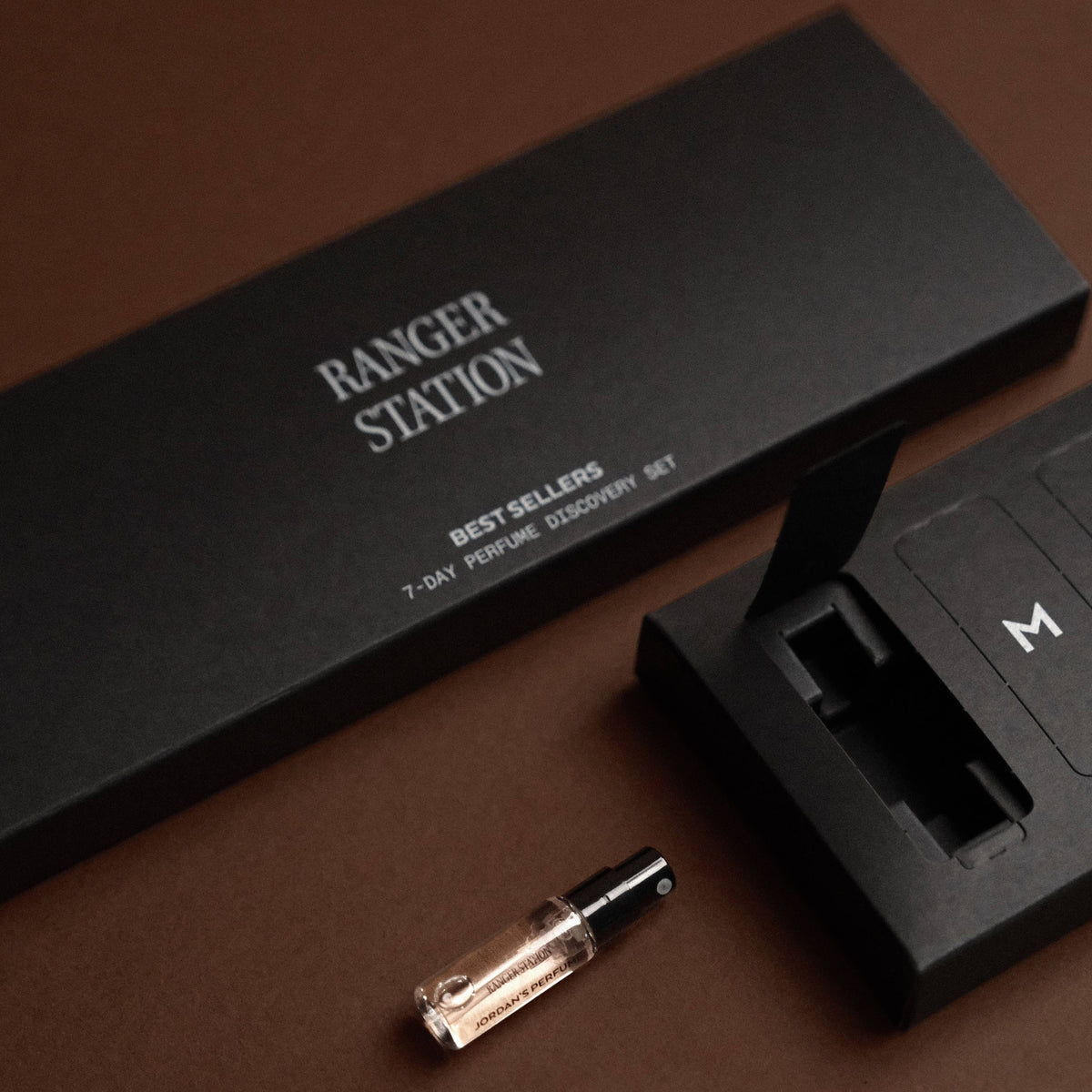 A small clear glass perfume vial with a black cap lies on a dark brown surface beside a black Ranger Station branded box and a black numbered tray.
