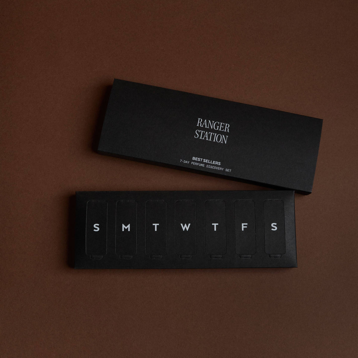 A black weekly planner style package with the days of the week labeled SMTWTFS, shown with its matching black lid or box labeled RANGER STATION, on a solid dark brown background.