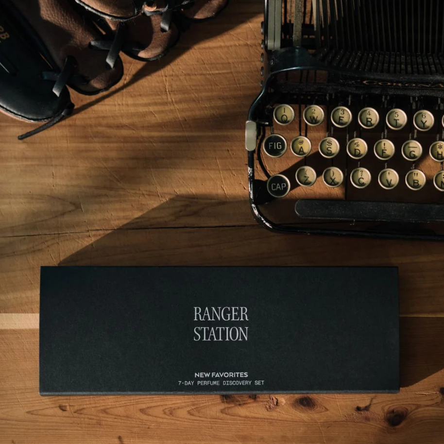 A dark rectangular perfume box labeled 'RANGER STATION' and 'NEW FAVORITES' sits on a wooden tabletop beside an open vintage typewriter and a pair of brown leather shoes.