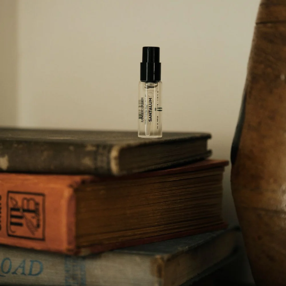 A small clear glass Ranger Station fragrance sample spray bottle with a black cap stands upright on top of a stack of vintage books against a warm beige background.