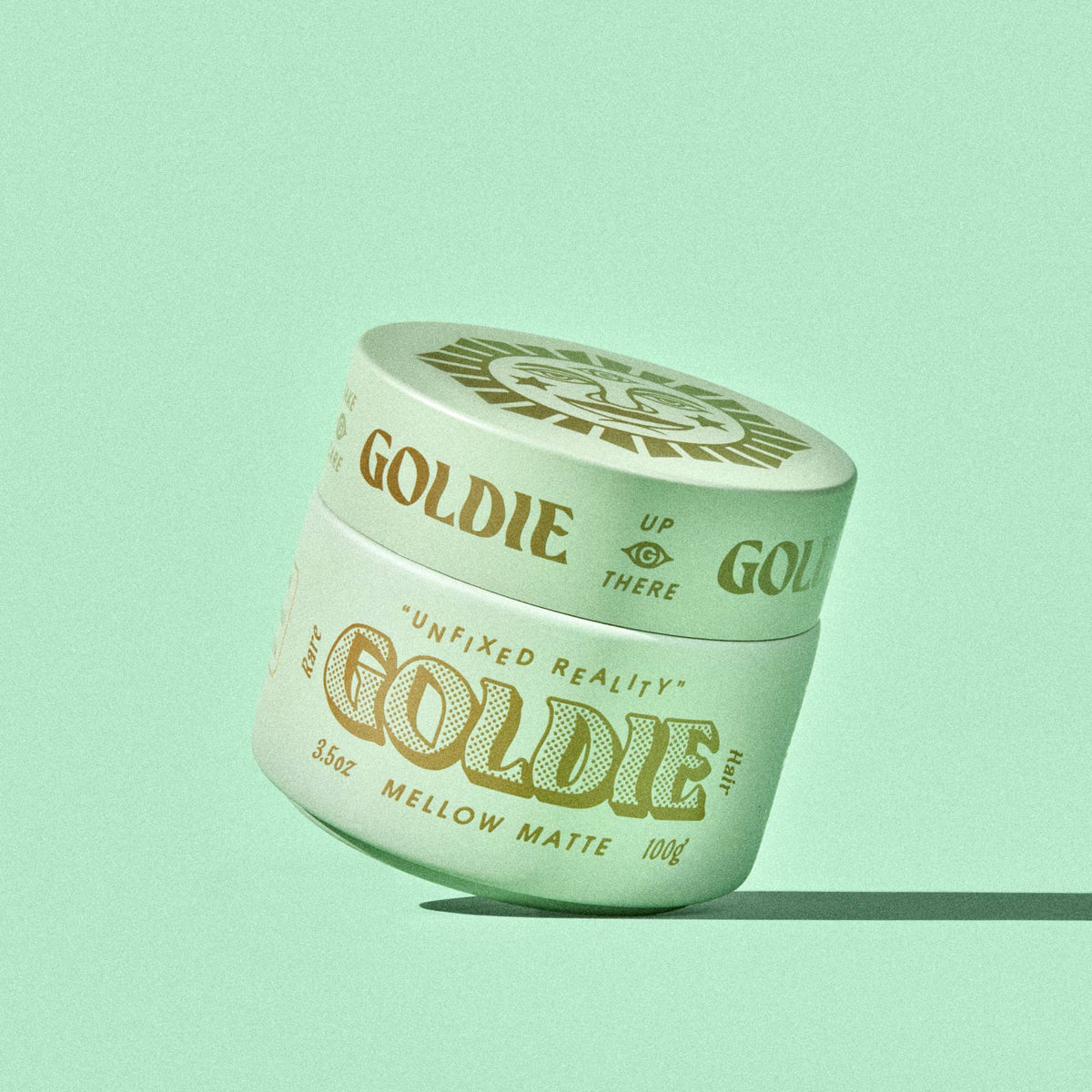 A single jar of Goldie Mellow Matte hair pomade sits at an angle on a solid mint green background. The jar is a pale mint green with gold lettering and a matching gold lid, casting a soft shadow to the right.