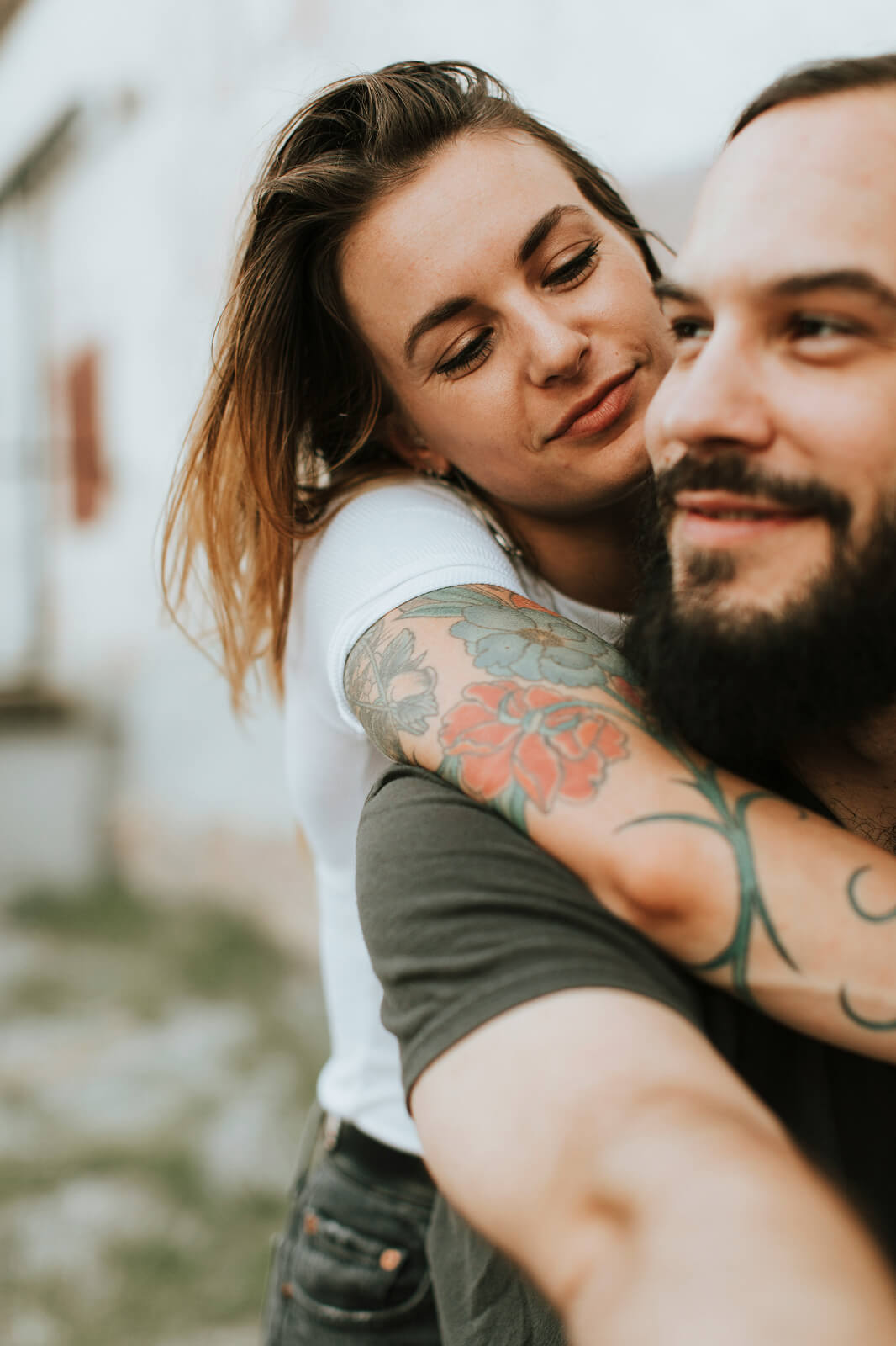 A close-up lifestyle shot of a couple embracing, with a woman with tattoos hugging a bearded man from behind.