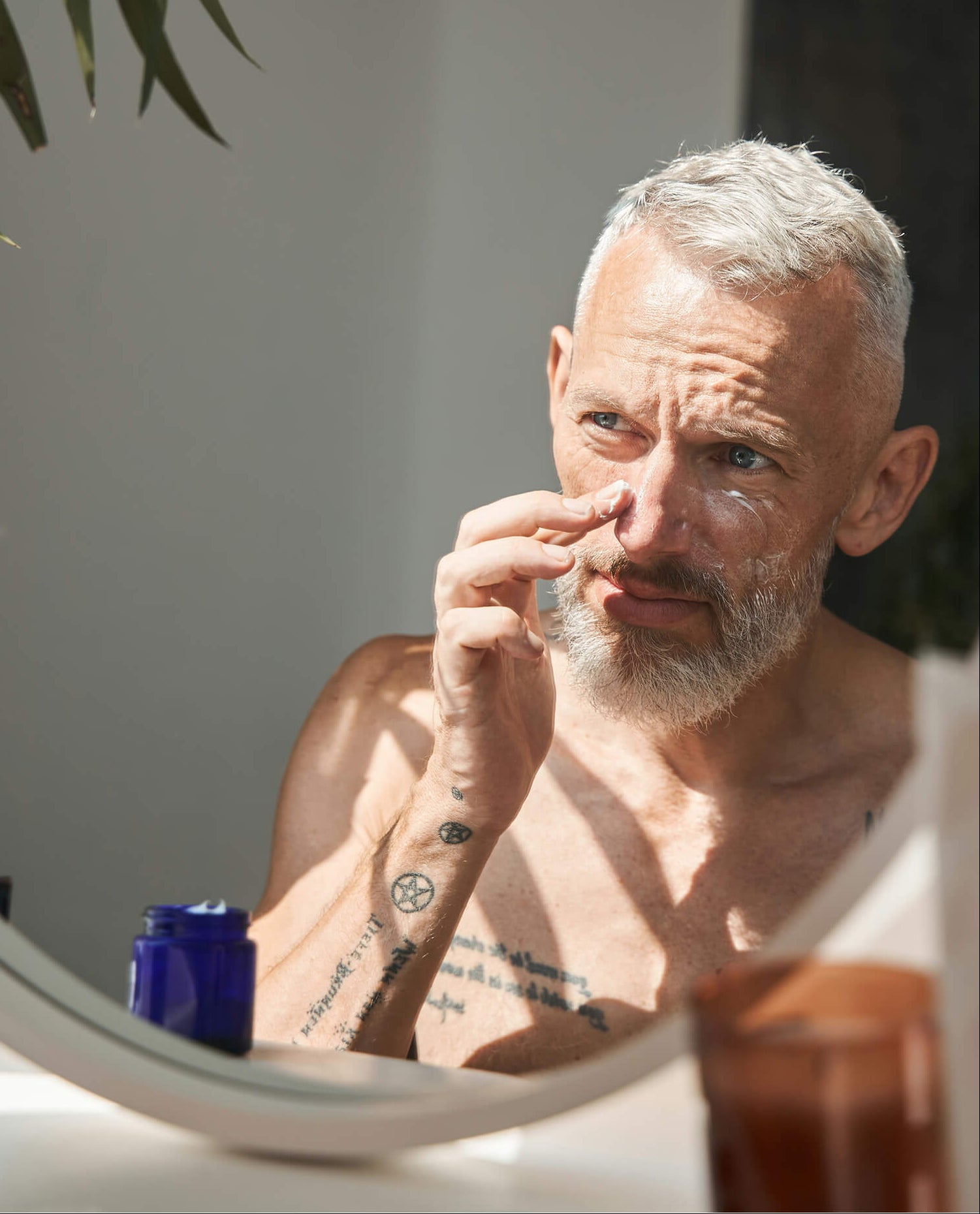 A man with short gray hair and a beard looks into a round mirror while touching his nose. He is shirtless, with a blue jar and a brown cup on the counter in front of him.